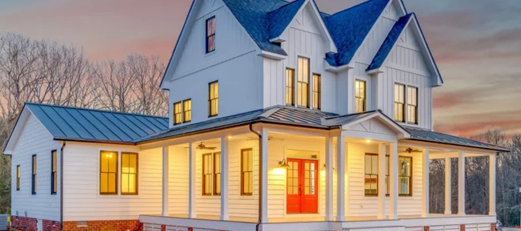 A two-story white farmhouse with board and batten siding, a wraparound porch, and a bright red front door at sunset