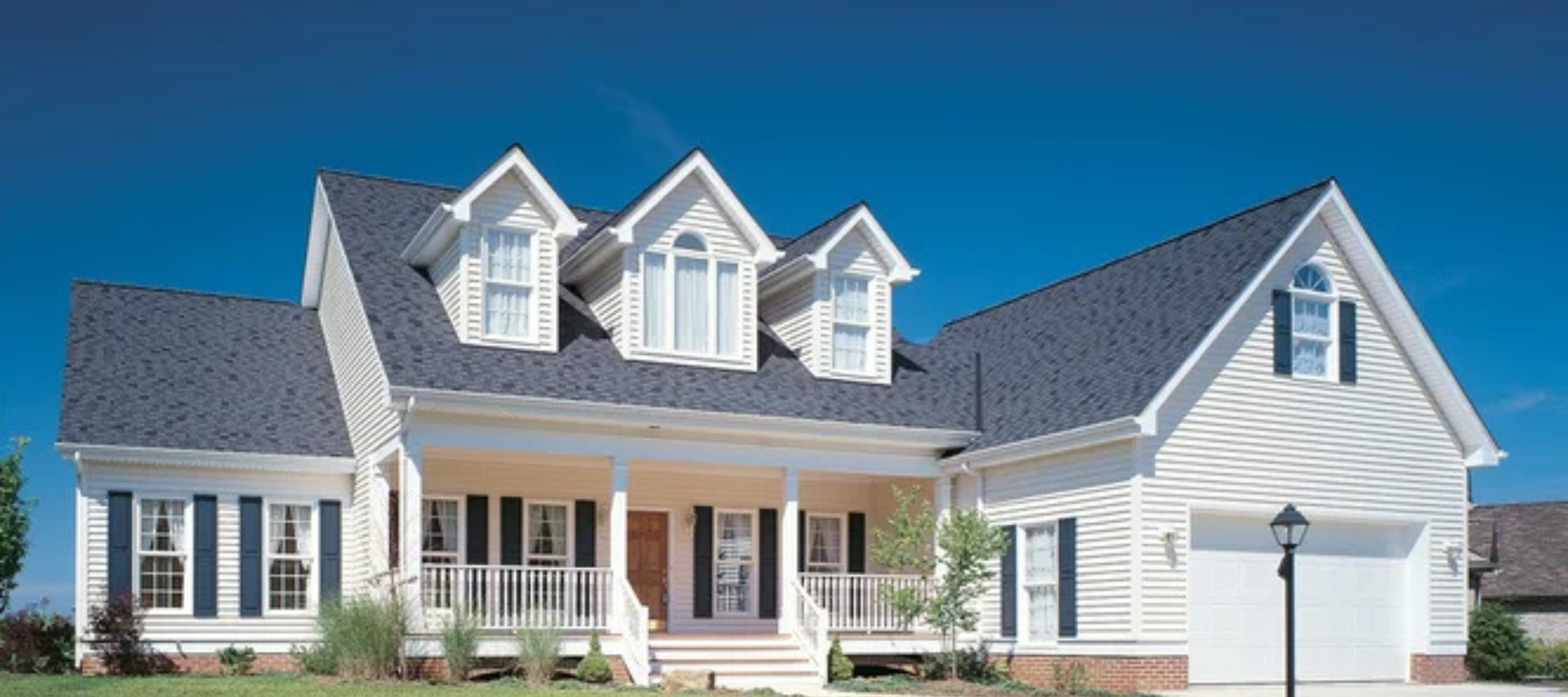 Large white home with dark shutters and dormer windows on a steep roof, sitting under a vibrant blue sky