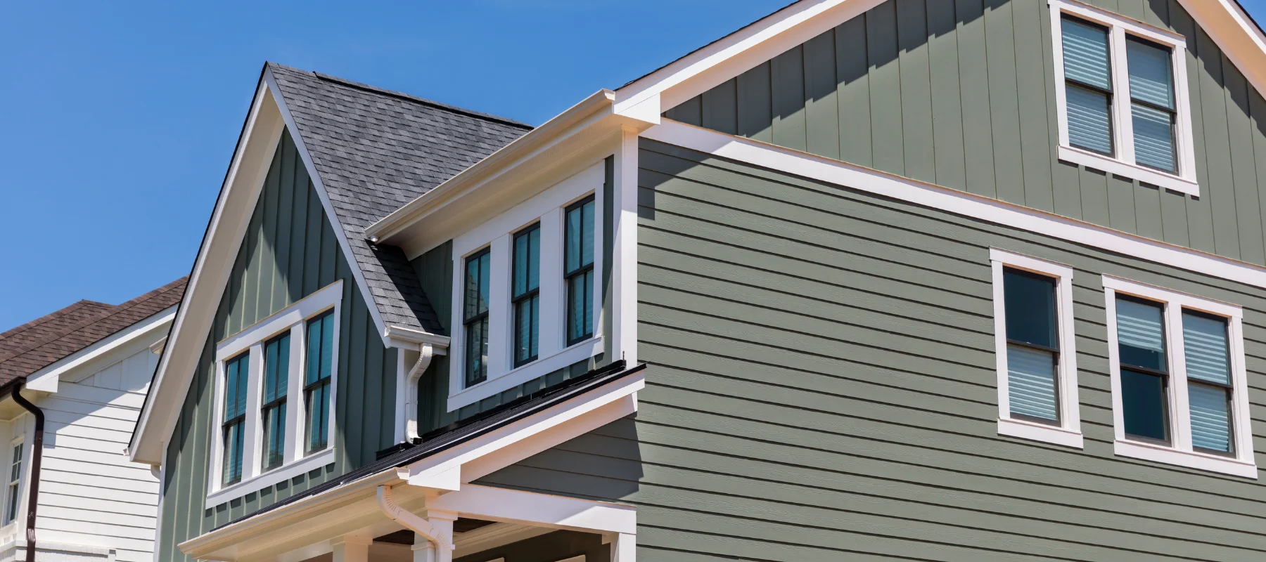 Two-story green home featuring a mix of horizontal and vertical siding, with white trim and multiple windows under a clear blue sky
