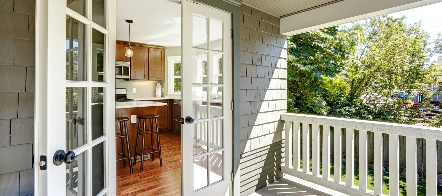 Open French doors lead from a kitchen to a shaded balcony with a wooden railing and green exterior siding