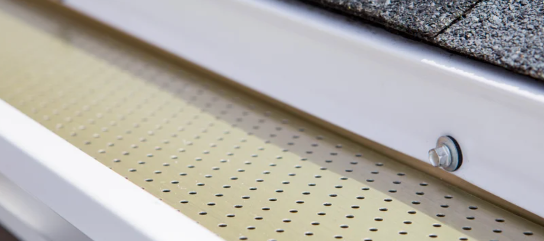 Macro view of a tan metal gutter guard with drainage holes installed beneath a white roof edge
