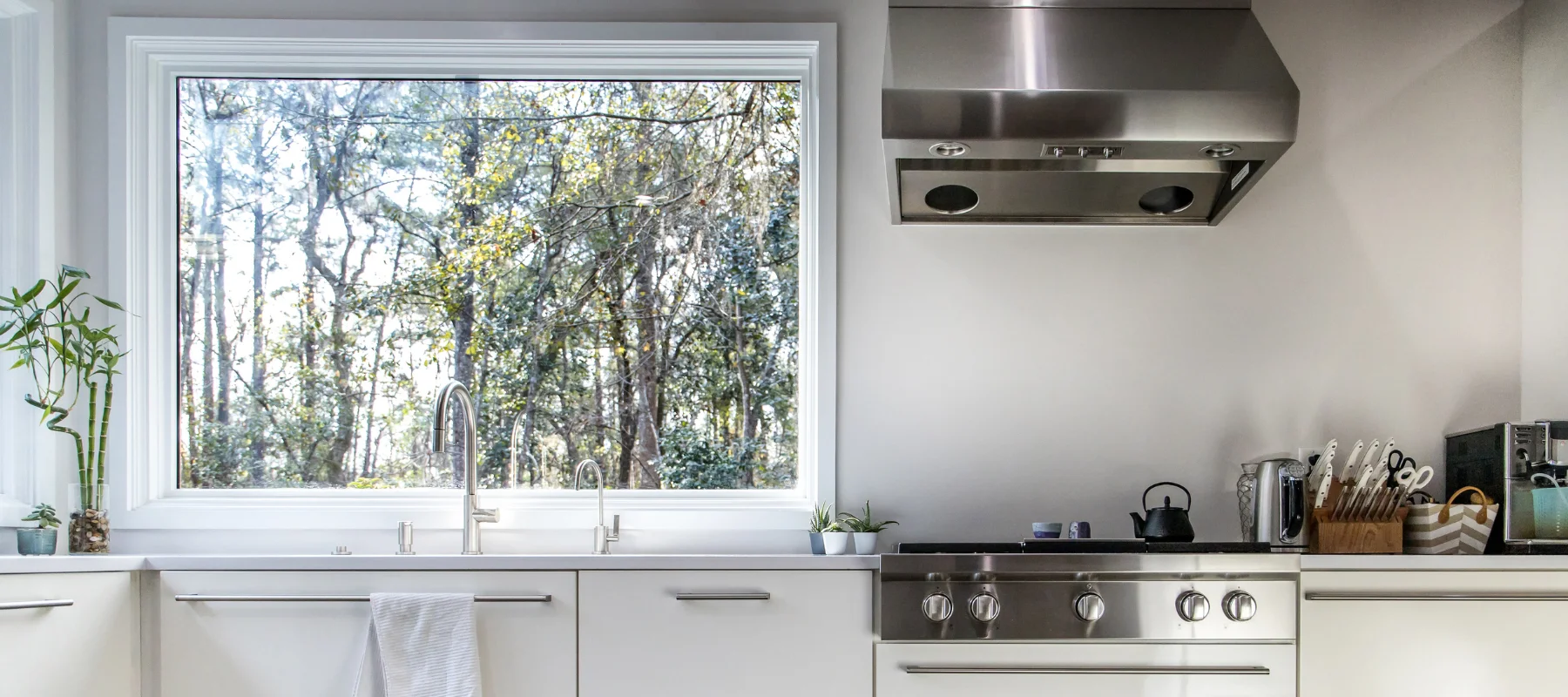 Large picture window above a modern white kitchen sink offering a clear view of the wooded backyard