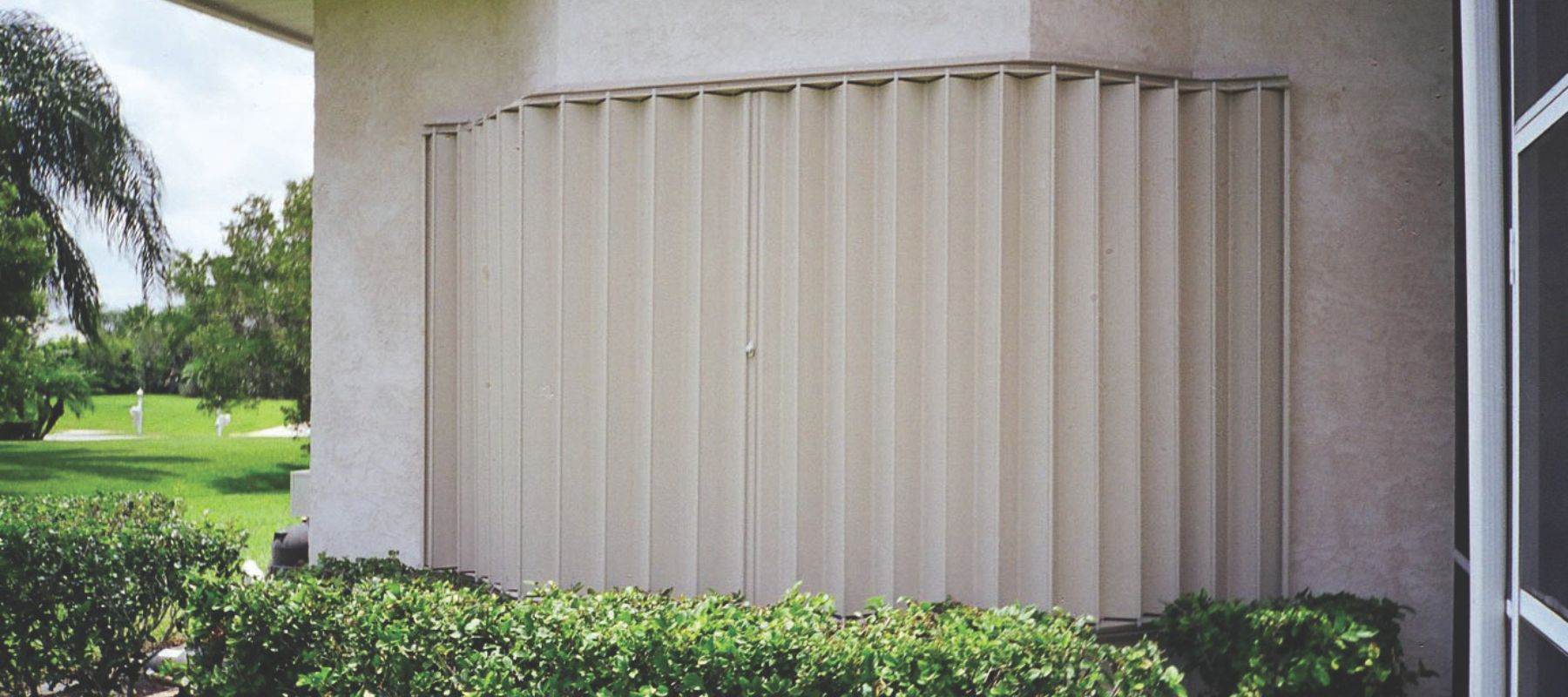Closed beige accordion-style storm shutters cover a large window on a stucco home