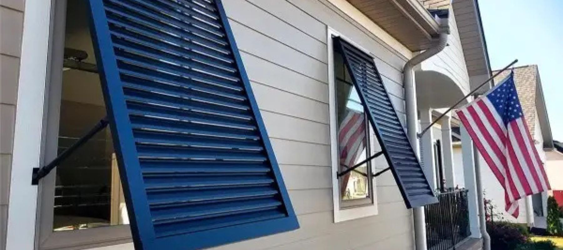 Exterior view of blue Bahama shutters propped open on a home with an American flag nearby