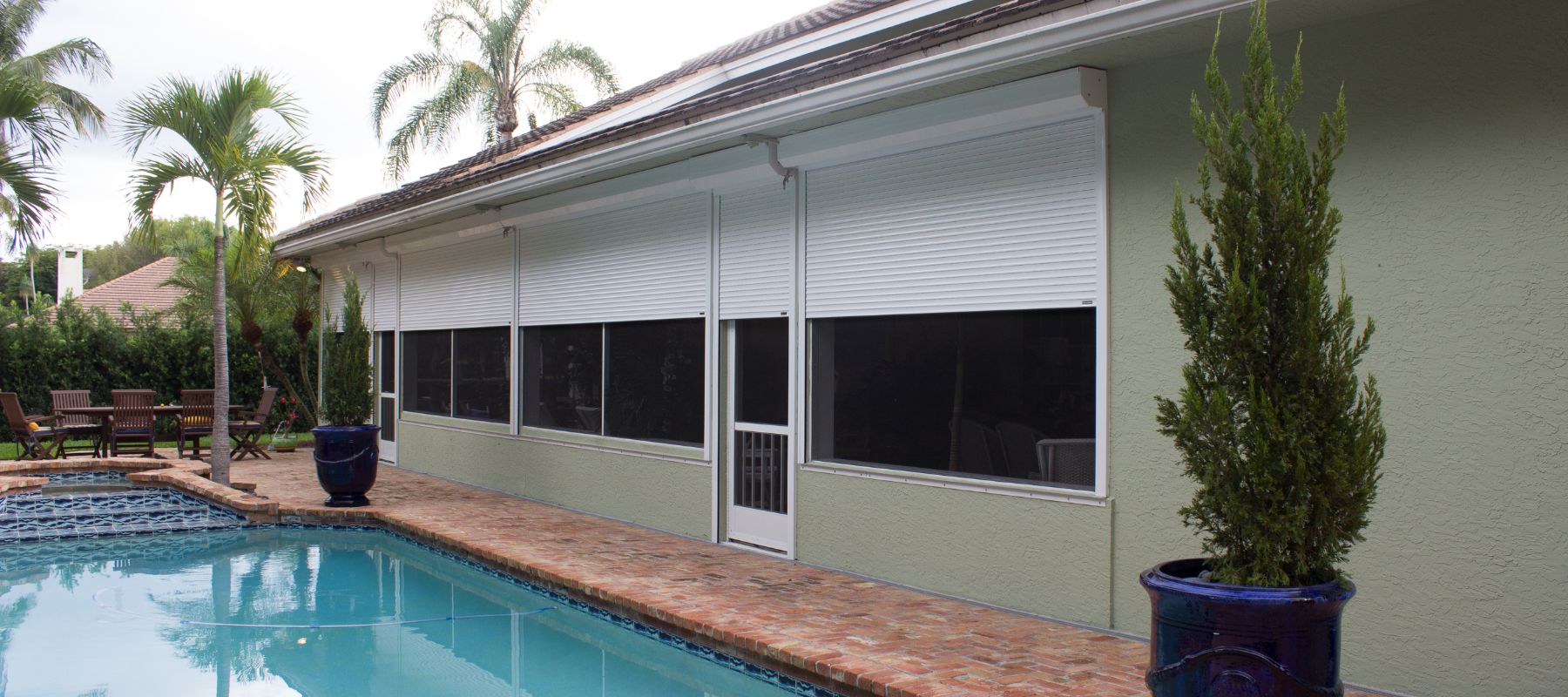 White roll-down storm shutters covering windows on the back of a green home near a pool