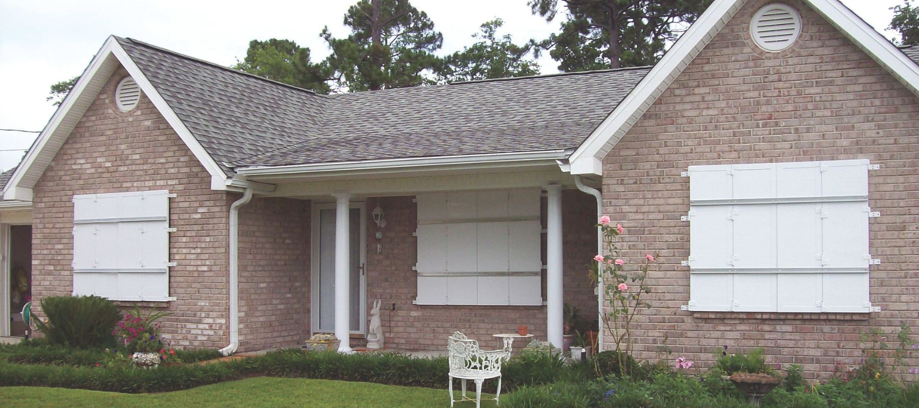 White metal storm panels are installed over the windows of a brick house with a covered front porch