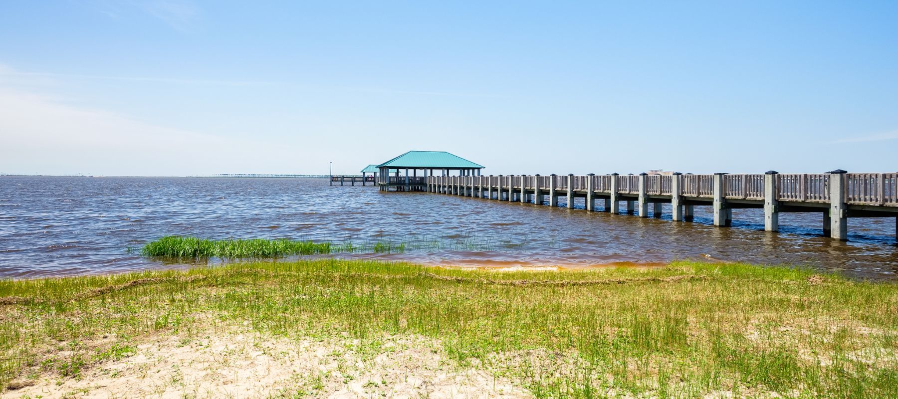 A long wooden pier with a green-roofed pavilion extending over calm water under a clear blue sky