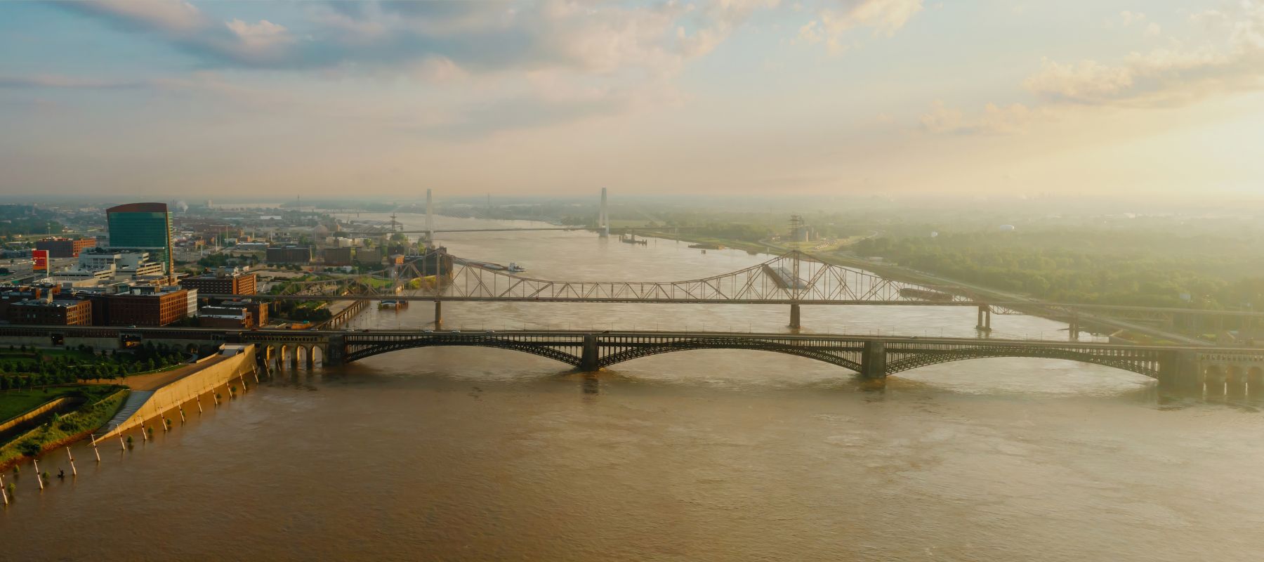 An aerial view captures multiple bridges spanning a wide river, with a cityscape on the left and a golden sunset haze on the right