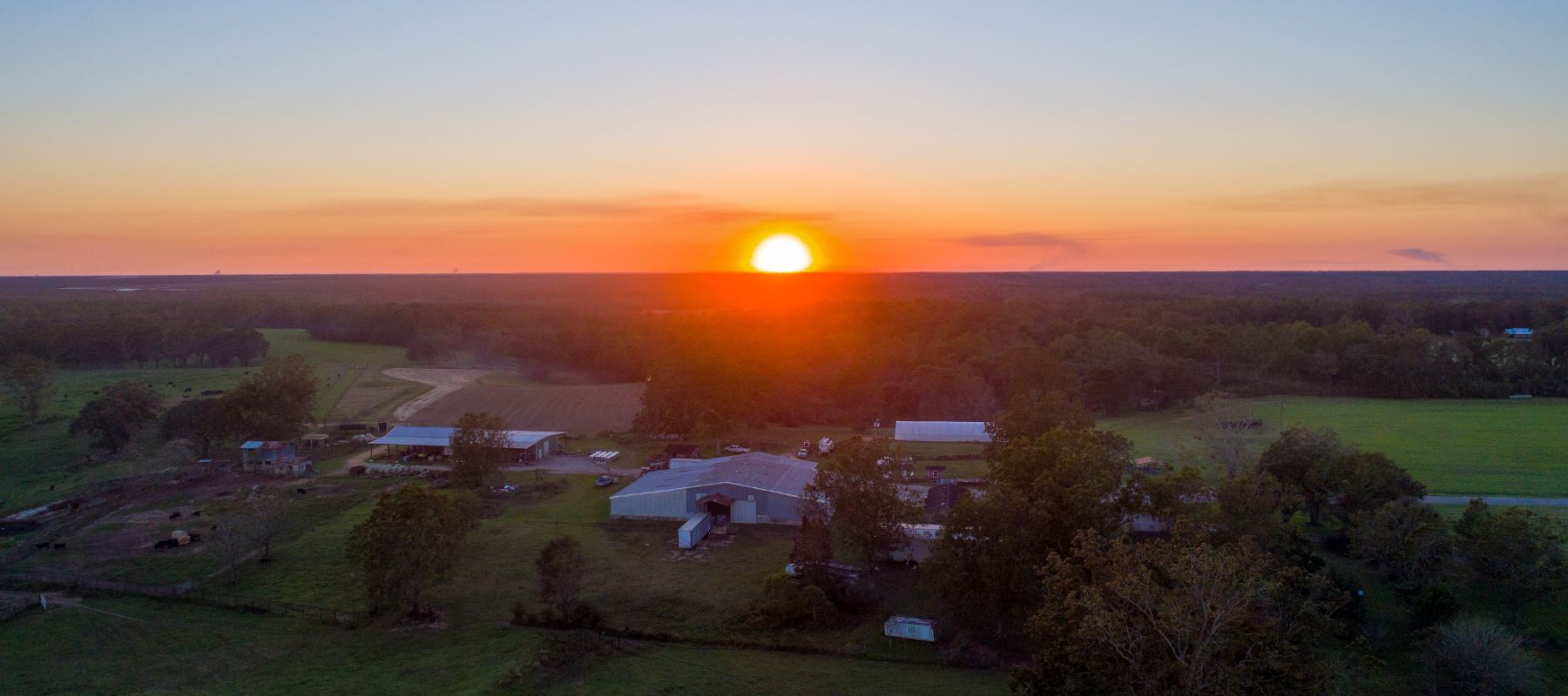 A bright orange sunset illuminates the horizon over a vast rural landscape with scattered farm buildings and dense trees