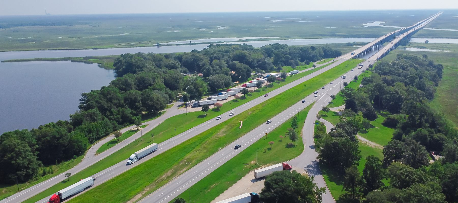 An aerial view shows a multi-lane highway stretching across a landscape of water and green trees, with a long bridge extending into the distance