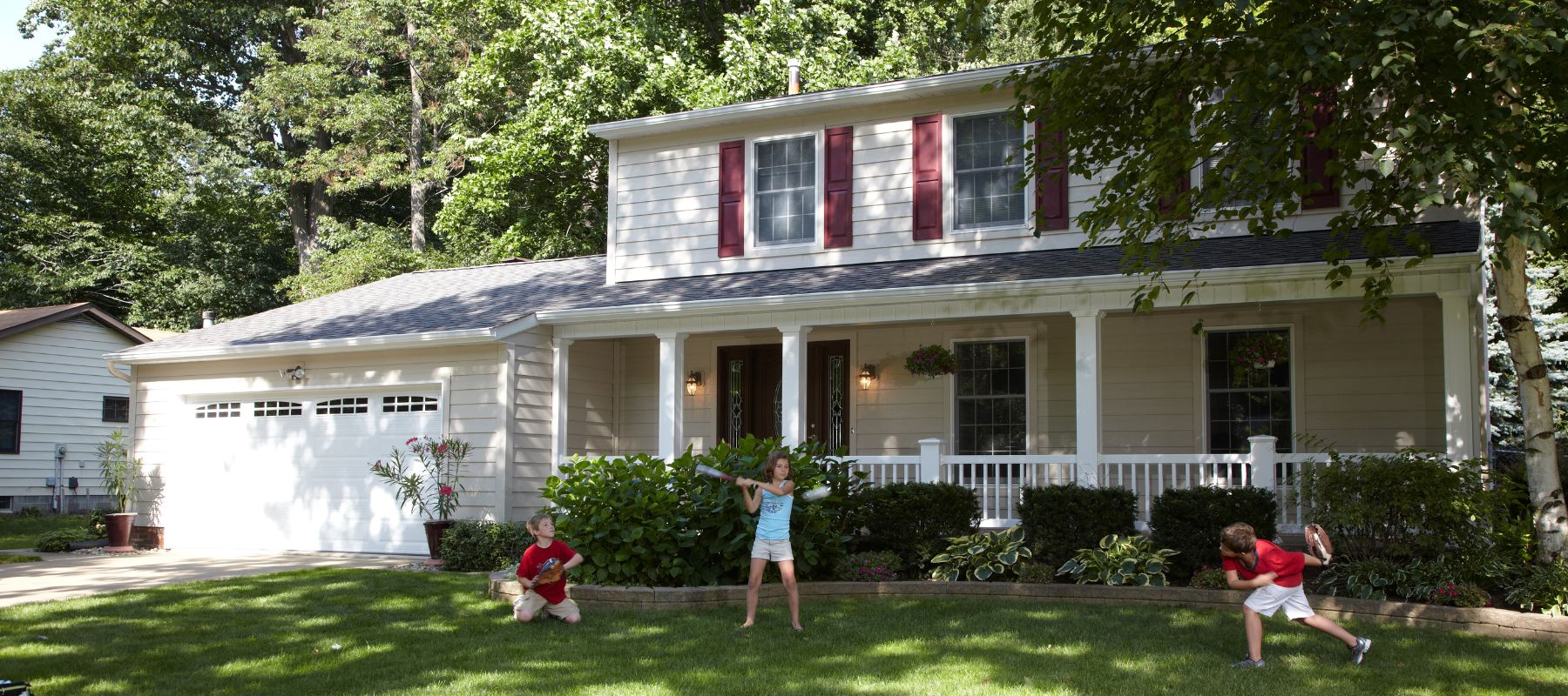 Three children play baseball in the front yard of a two-story beige house with red shutters and a wide porch