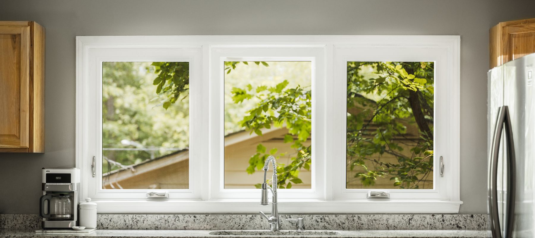 A kitchen window with three panes looks out onto green trees and a partial view of a house, above a sink with a faucet and a granite countertop