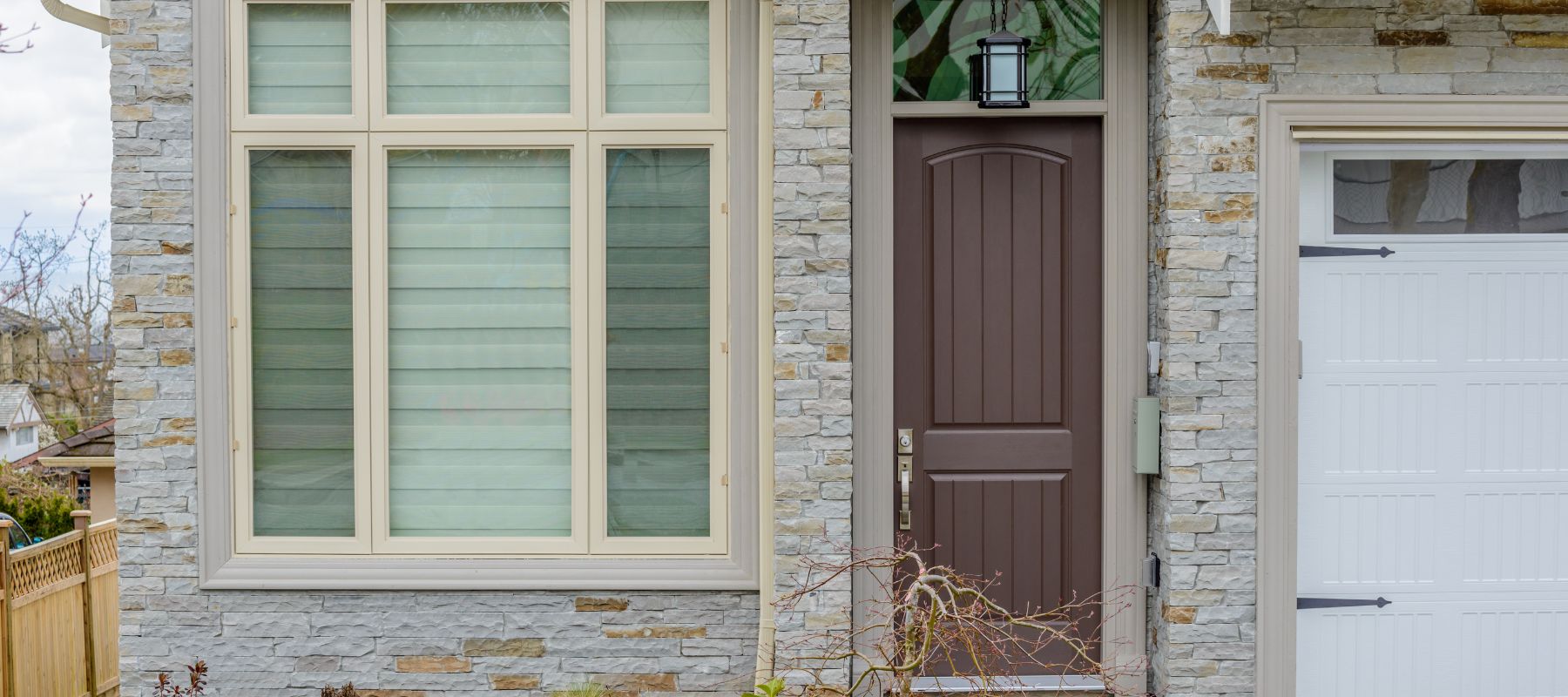 The front of a house features light-colored stone siding, a brown front door with a lantern, and a large window with tan frames and blinds