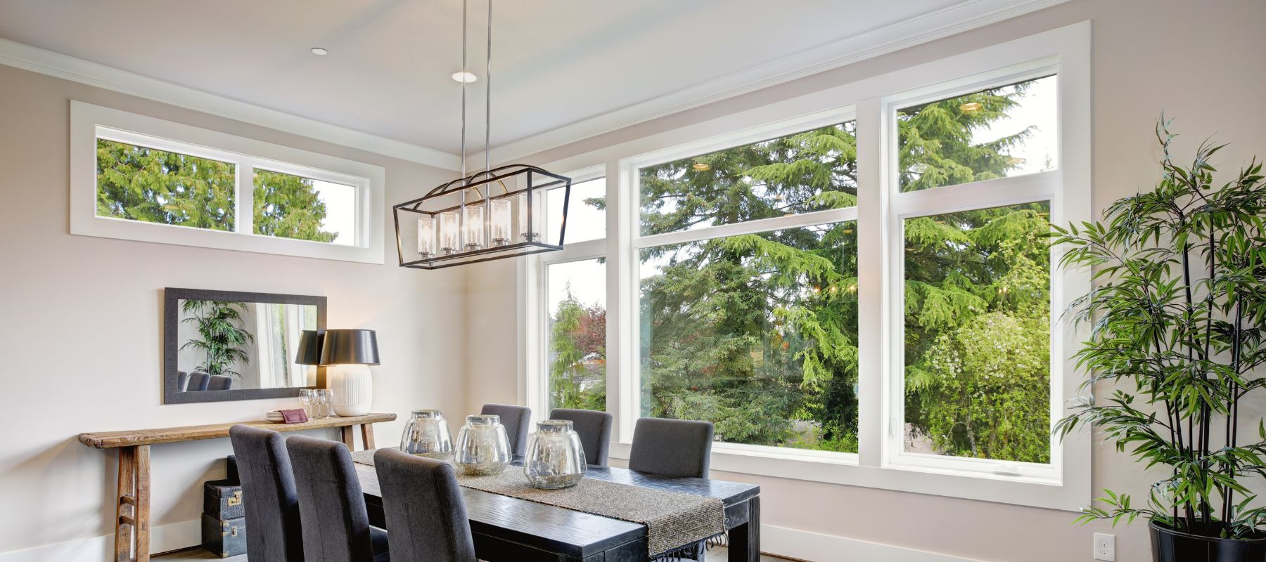 A bright dining room features a long dark dining table surrounded by chairs, large windows looking out onto green trees, and a modern chandelier