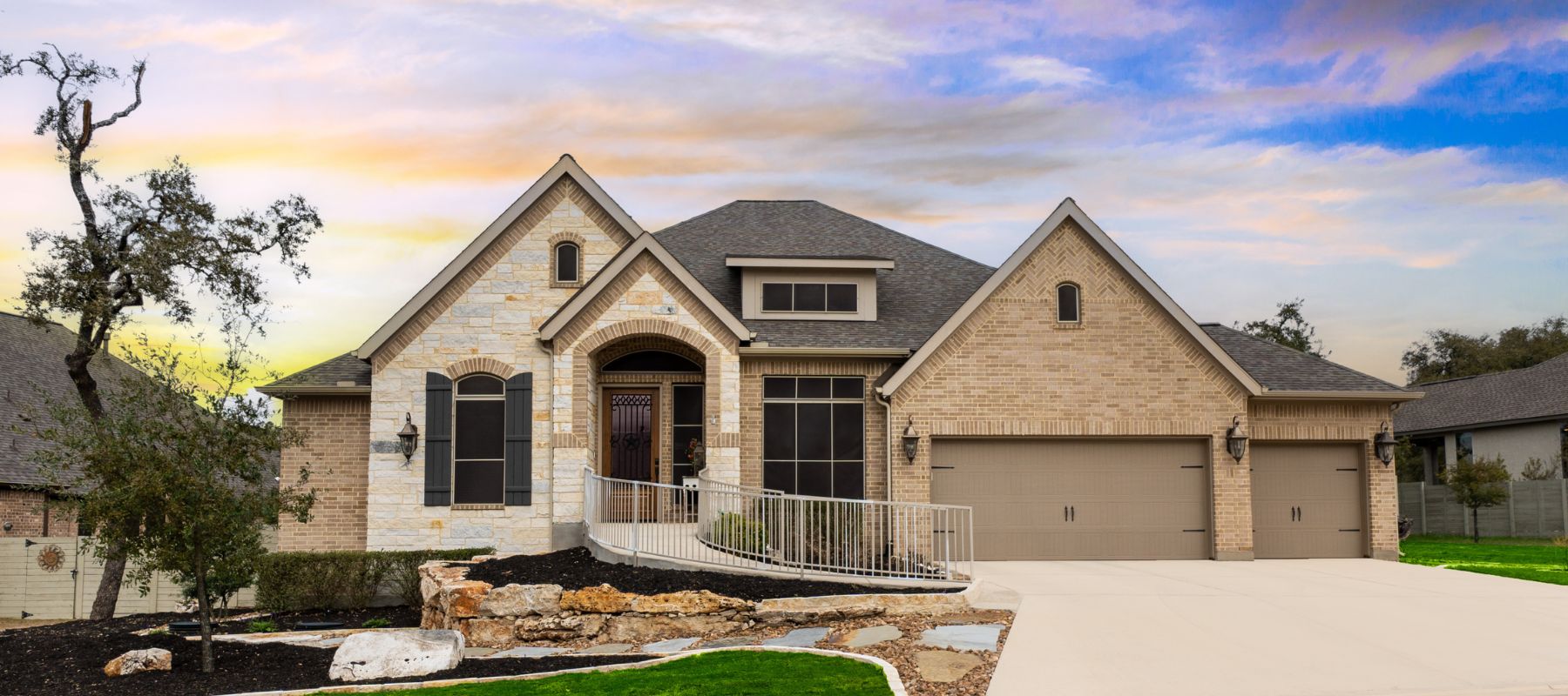 A large, elegant suburban home with a mix of brick and stone siding features multiple gables, a two-car garage, and a welcoming entrance with a ramp, under a colorful sky