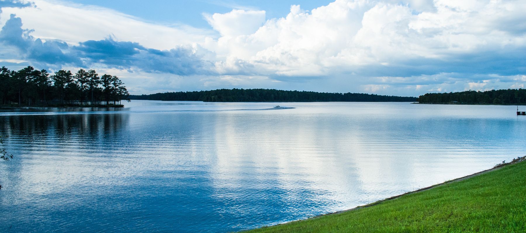 A wide, serene lake reflects the cloudy sky, with dark green trees lining the distant shores and a small boat leaving a wake